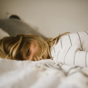 Person mit blondem Haar liegt bäuchlings auf einem Bett, den Kopf zur Seite gedreht. Sie trägt ein weißes Shirt mit schwarzen Streifen. Im Hintergrund sind neutrale Kissen und Bettwäsche zu sehen.