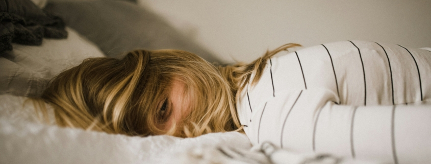 Person mit blondem Haar liegt bäuchlings auf einem Bett, den Kopf zur Seite gedreht. Sie trägt ein weißes Shirt mit schwarzen Streifen. Im Hintergrund sind neutrale Kissen und Bettwäsche zu sehen.