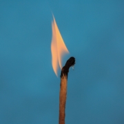 So wirken Impacttechniken Nahaufnahme eines brennenden Streichholzes vor einem einfarbig blauen Hintergrund. Die Flamme ist gelb-orange und leicht nach links geneigt, während der obere Teil des Streichholzes schwarz verkohlt ist.