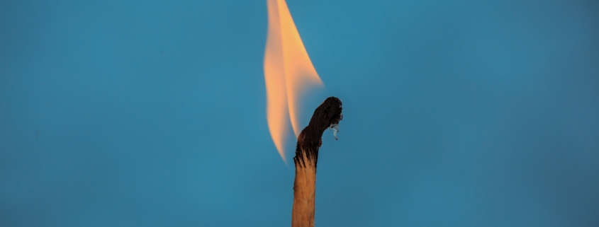 Nahaufnahme eines brennenden Streichholzes vor einem einfarbig blauen Hintergrund. Die Flamme ist gelb-orange und leicht nach links geneigt, während der obere Teil des Streichholzes schwarz verkohlt ist.